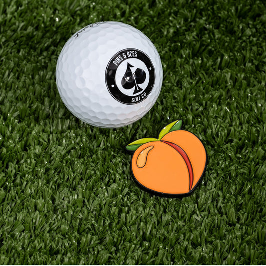 A Pins and Aces golf ball rests on artificial grass beside a Peach Ball Marker shaped like a cartoon peach.