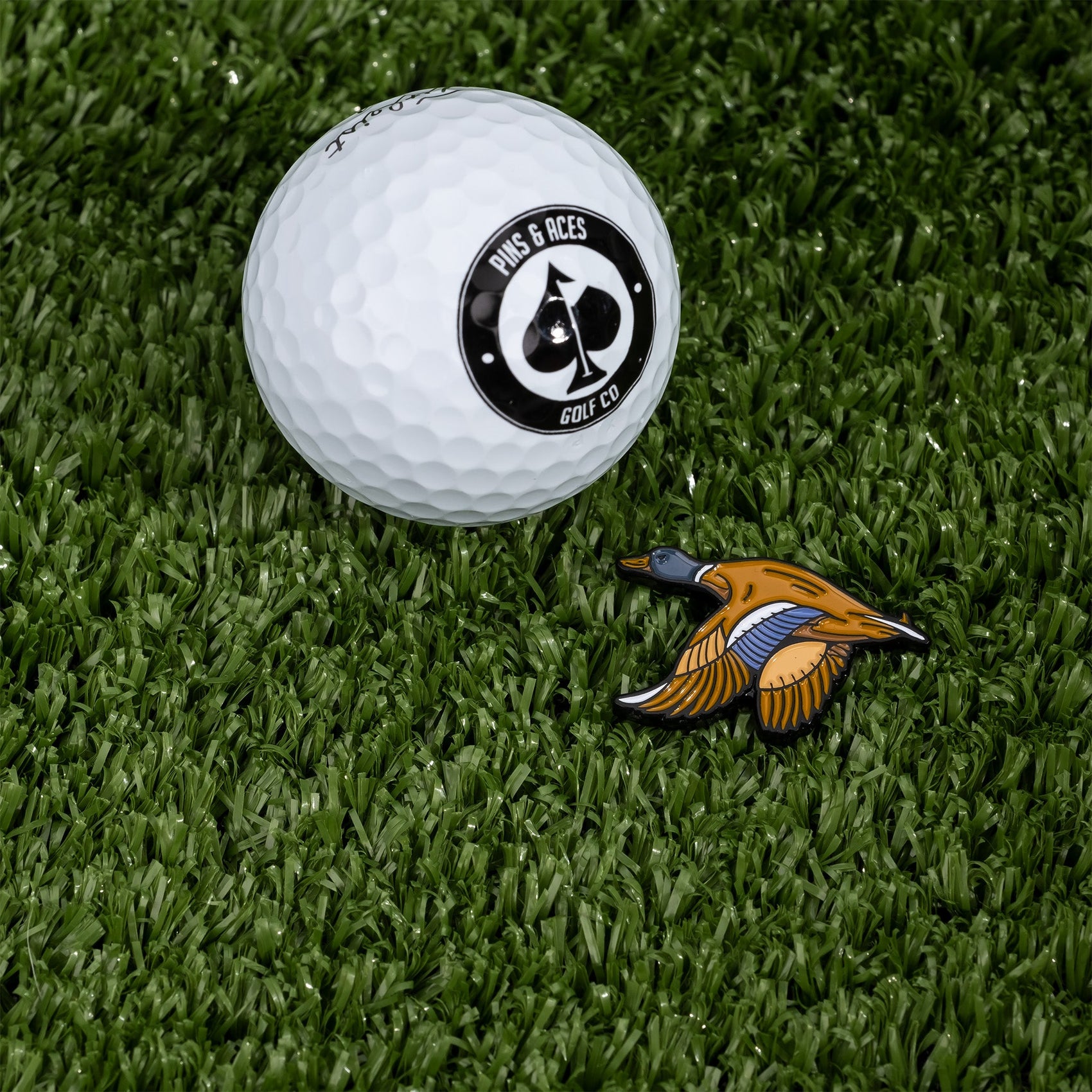 A Pins and Aces golf ball and the Flying Mallard Ball Marker rest on green artificial turf.