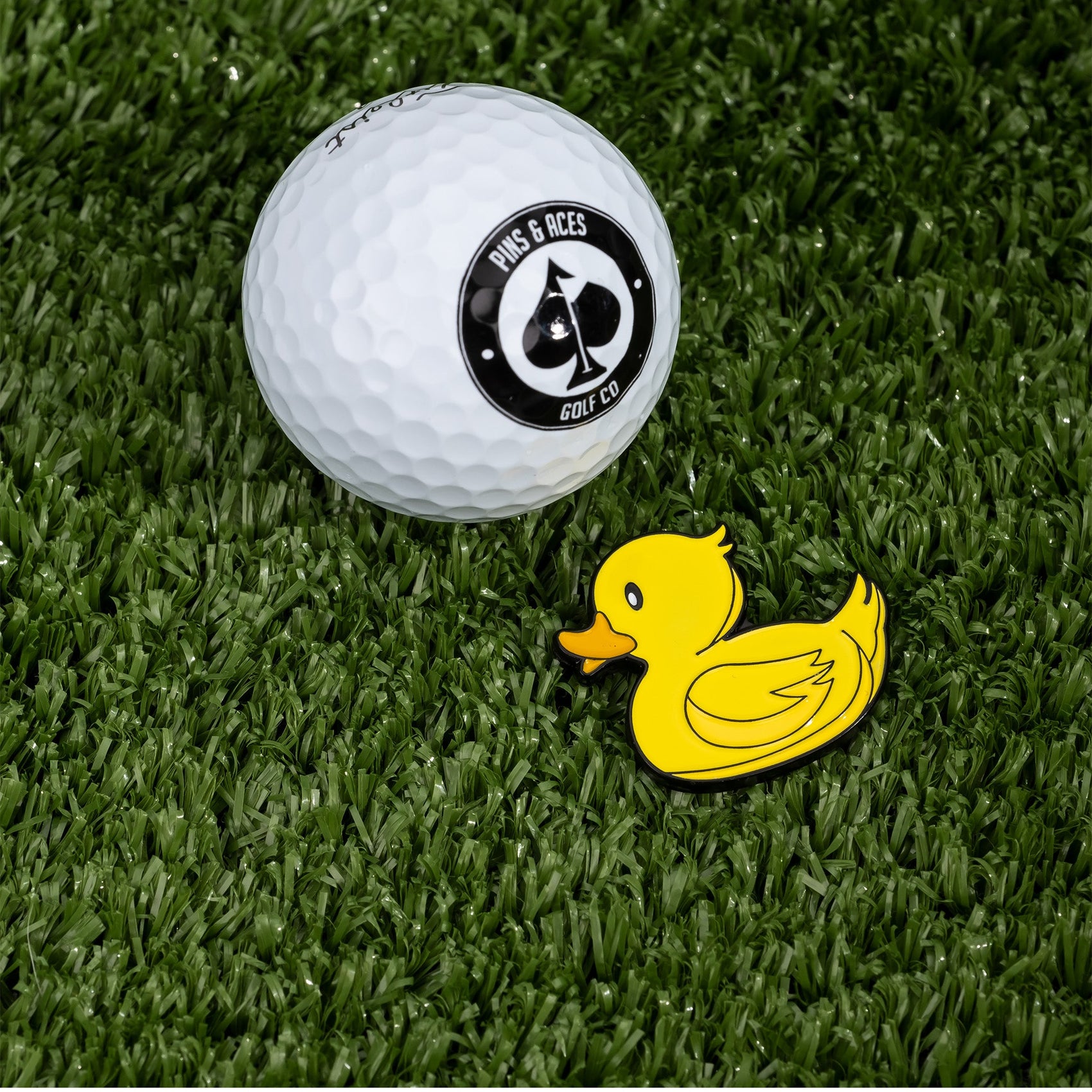 A white golf ball with a black Pins and Aces logo rests on grass beside the Rubber Ducky Ball Marker, a small yellow enamel marker shaped like a rubber duck.