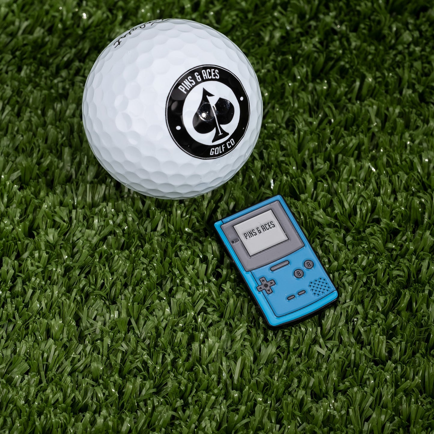 A white golf ball with a Pins and Aces logo rests on green turf beside the Game Boy Ball Marker, a blue, retro gaming-inspired golf accessory from Pins and Aces, featuring "PINS & ACES" displayed on its screen.