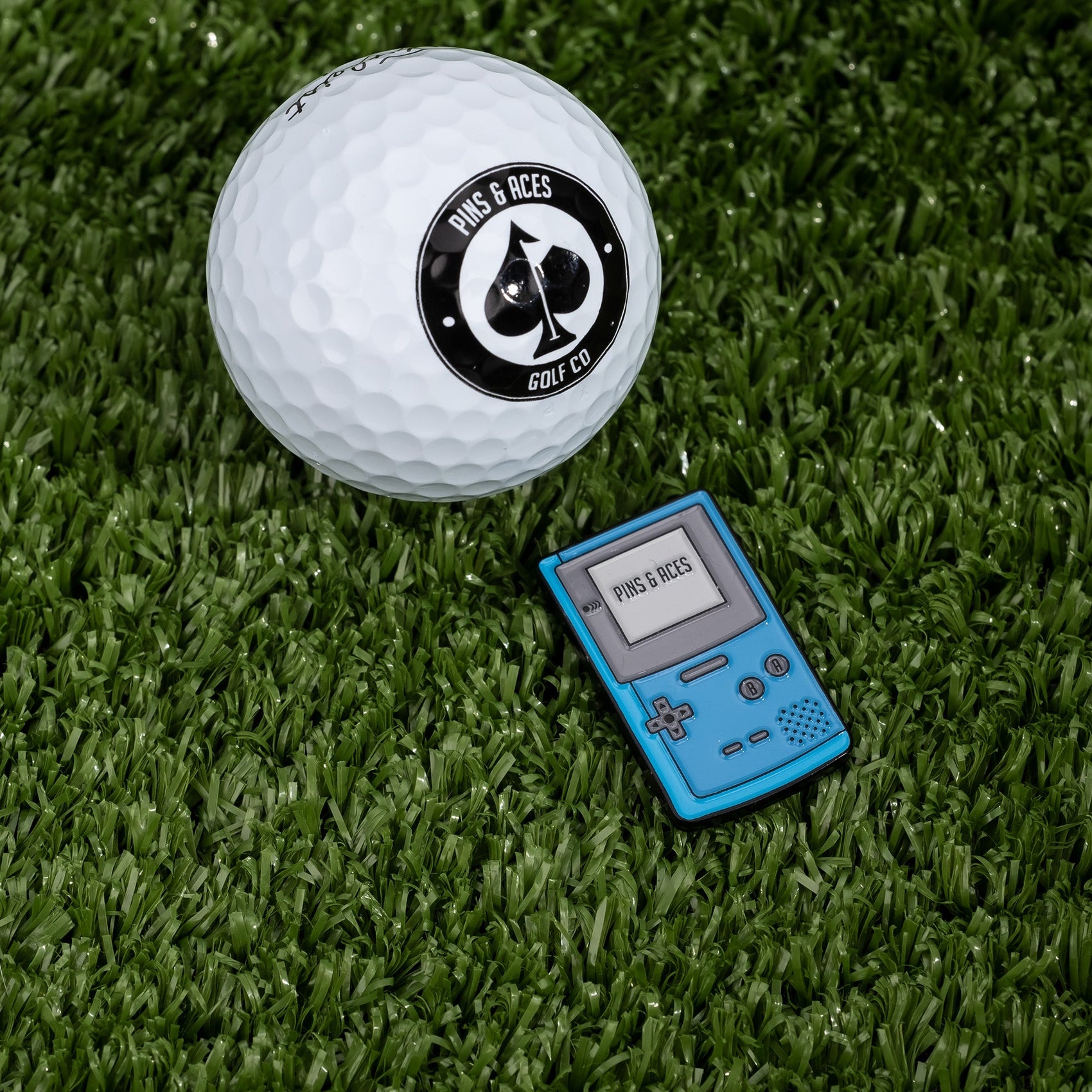 A white golf ball with a Pins and Aces logo rests on green turf beside the Game Boy Ball Marker, a blue, retro gaming-inspired golf accessory from Pins and Aces, featuring "PINS & ACES" displayed on its screen.
