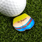 A golf ball and divot tool rest on artificial grass beside Logie Joe's Golf "No Ragrets" pin, featuring a cartoon lower back with a No Ragrets tattoo above blue pants and a red belt.