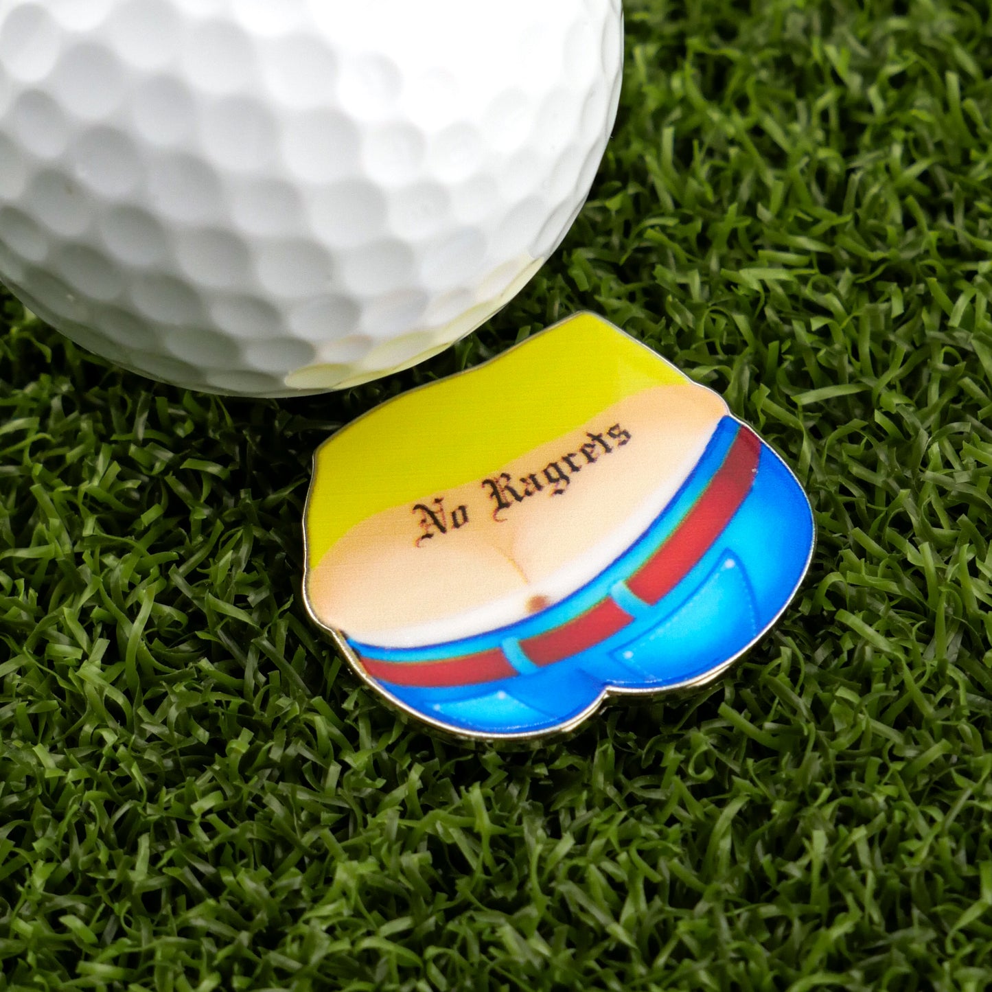 A golf ball and divot tool rest on artificial grass beside Logie Joe's Golf "No Ragrets" pin, featuring a cartoon lower back with a No Ragrets tattoo above blue pants and a red belt.