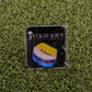An enamel pin on grass illustrates yellow shorts with red and blue accents plus "No Ragrets." The black card shows Logie Joe's Golf with a palm tree. Perfect for fans of unique, premium golf accessories.
