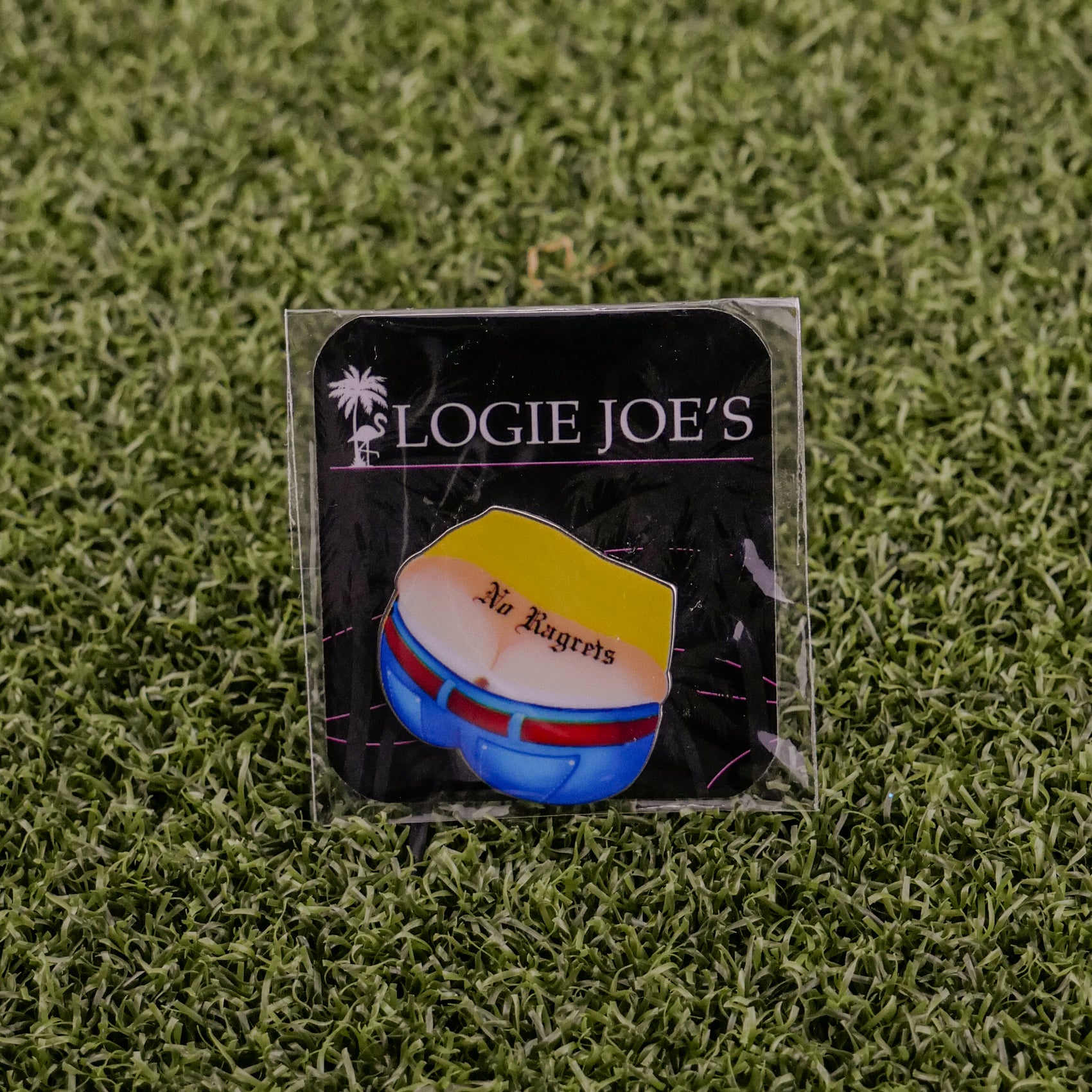 An enamel pin on grass illustrates yellow shorts with red and blue accents plus "No Ragrets." The black card shows Logie Joe's Golf with a palm tree. Perfect for fans of unique, premium golf accessories.