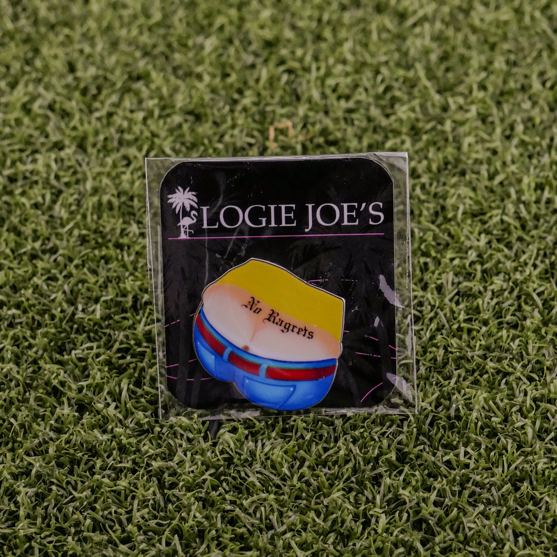 An enamel pin on grass illustrates yellow shorts with red and blue accents plus "No Ragrets." The black card shows Logie Joe's Golf with a palm tree. Perfect for fans of unique, premium golf accessories.