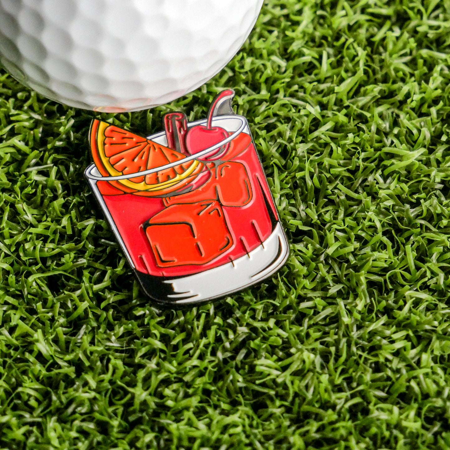 A golf ball on artificial grass sits beside the Ballmarkers.com Old Fashioned Ball Marker, a colorful enamel pin shaped like a cocktail with ice cubes, an orange slice, and a cherry garnish.