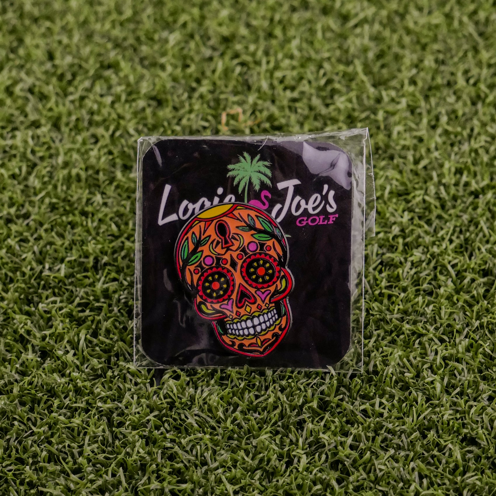 The Orange Sugar Skull ball marker by Logie Joe's Golf features a vibrant design and logo, packaged in plastic on green artificial grass—an ideal pick for fans of premium golf accessories.