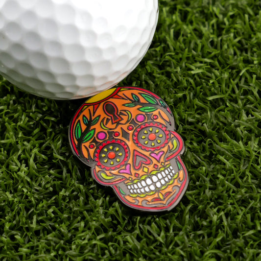 A golf ball sits on artificial grass next to the Ghouls of the Green by Logie Joe's Golf Orange Sugar Skull Ball Marker, a bold accessory featuring artistic orange, pink, and green patterns.