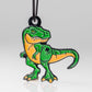 The Pins and Aces T-Rex Ornament is a keychain featuring a cartoon green and orange T-Rex with a fierce expression and bared teeth, hanging from a black loop on a white background.