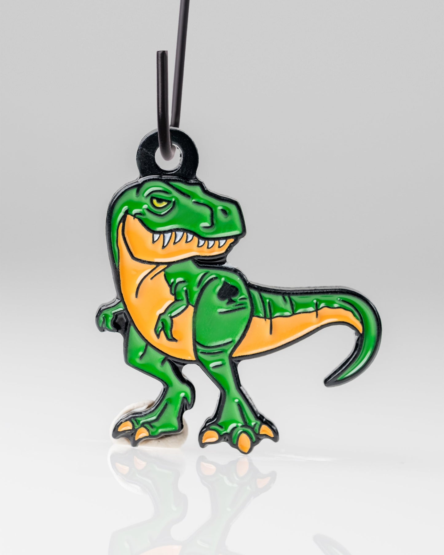 The Pins and Aces T-Rex Ornament is a keychain featuring a cartoon green and orange T-Rex with a fierce expression and bared teeth, hanging from a black loop on a white background.