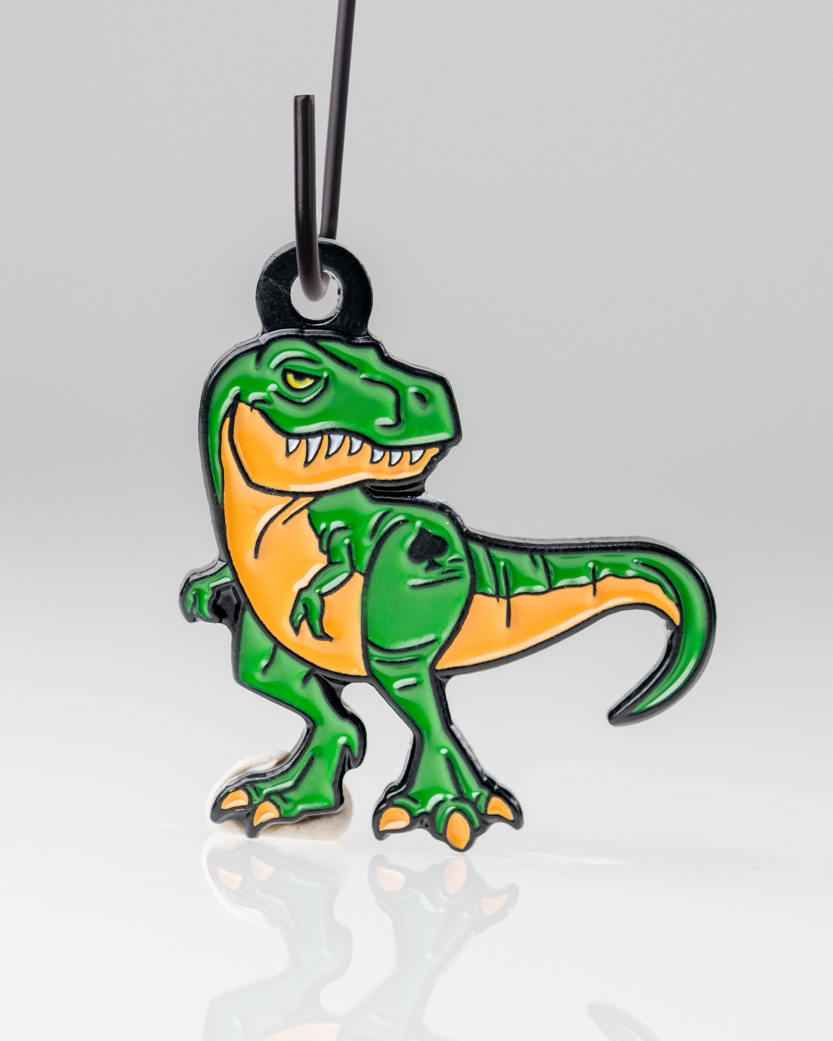 The Pins and Aces T-Rex Ornament is a keychain featuring a cartoon green and orange T-Rex with a fierce expression and bared teeth, hanging from a black loop on a white background.