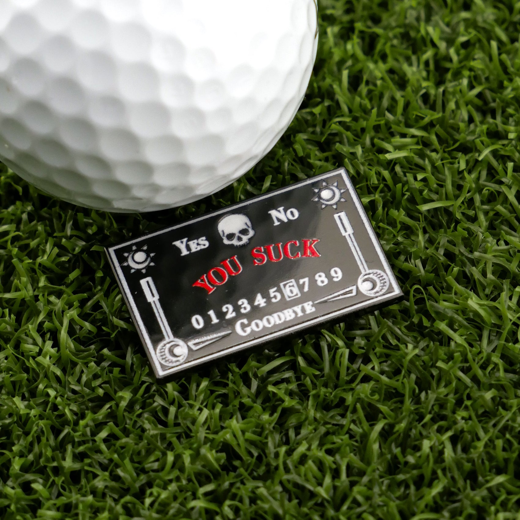 A golf ball rests on artificial grass beside Logie Joe's Golf Ouija Board ball marker—a hilarious accessory styled like a Ouija board, featuring bold red YOU SUCK text with Yes/No options and Goodbye at the bottom. Perfect for fun on the course!.