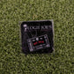 A black Logie Joe's Golf Ouija Board ball marker sits on artificial grass, featuring YOU SUCK in red, YES, NO, 0-9, and GOODBYE—making it a unique and funny golf accessory.