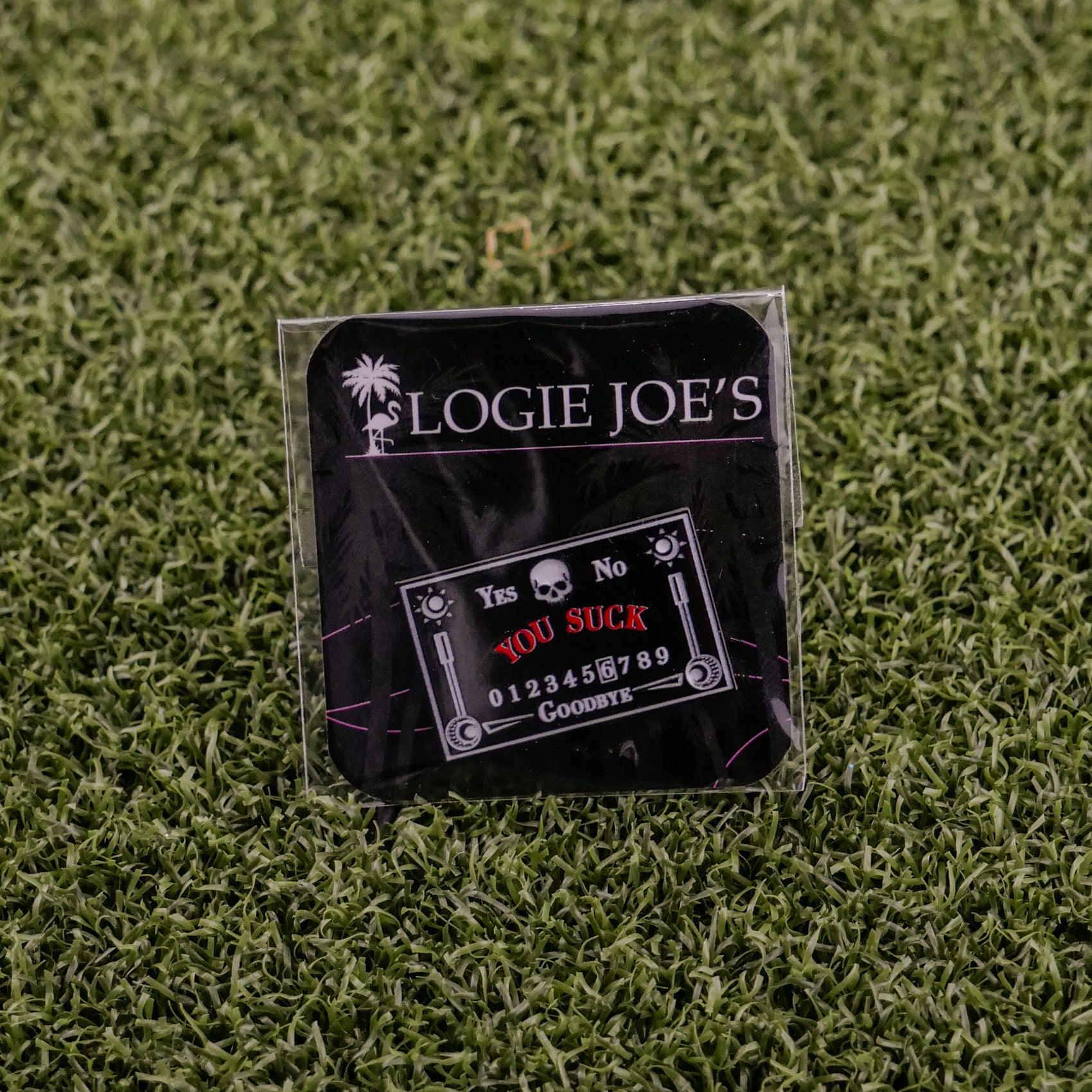 A black Logie Joe's Golf Ouija Board ball marker sits on artificial grass, featuring YOU SUCK in red, YES, NO, 0-9, and GOODBYE—making it a unique and funny golf accessory.