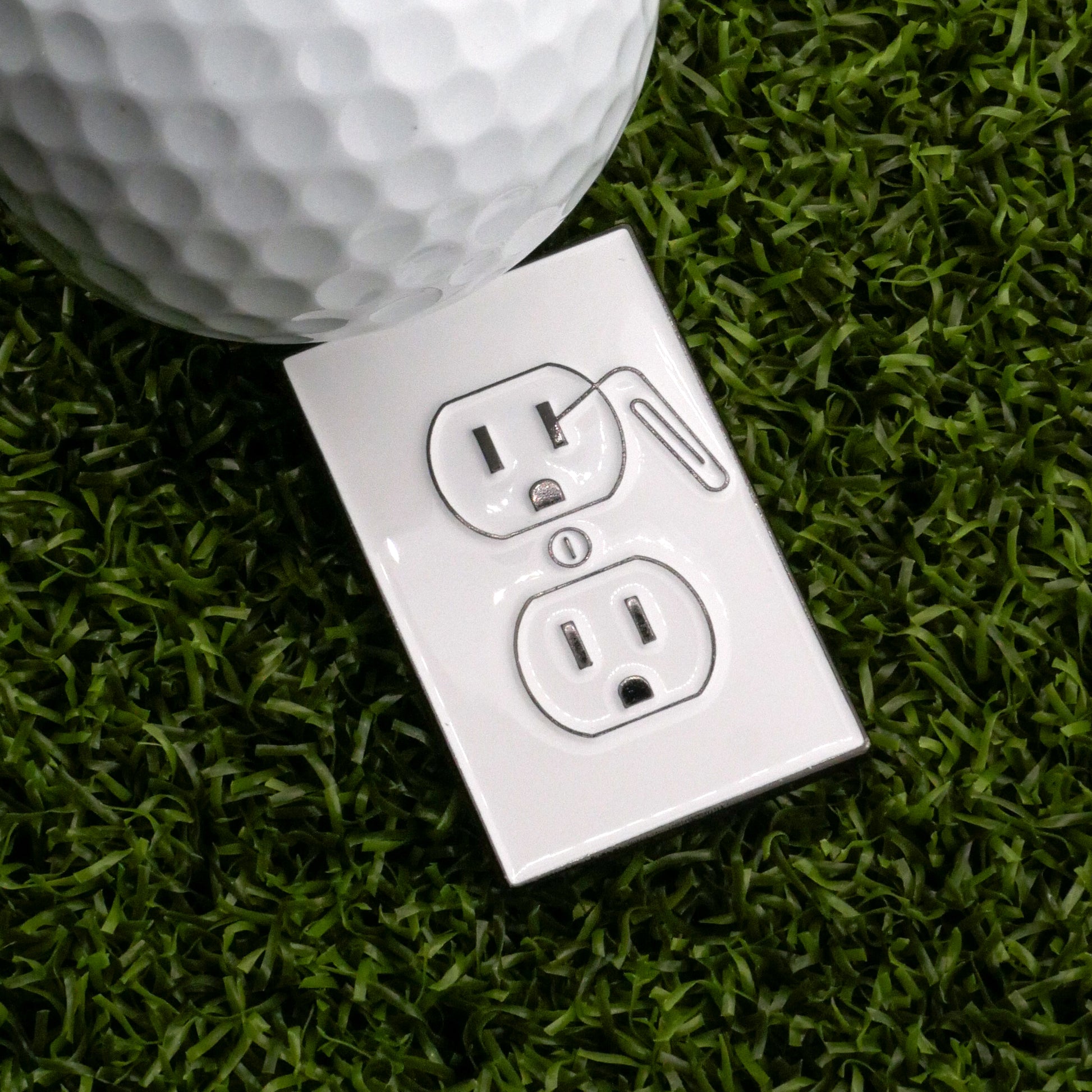 The Shocking Outlet Ball Marker by Ballmarkers.com is a white outlet cover with a golf club and ball design on green artificial grass, paired with a real golf ball that can double as a unique marker.