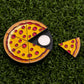 A Logie Joe's Golf Pepperoni Pizza "Slice" Premium Ball Marker, featuring a pizza with yellow cheese, red pepperoni, and one slice removed to reveal the silver ball marker, is displayed on a grassy surface.