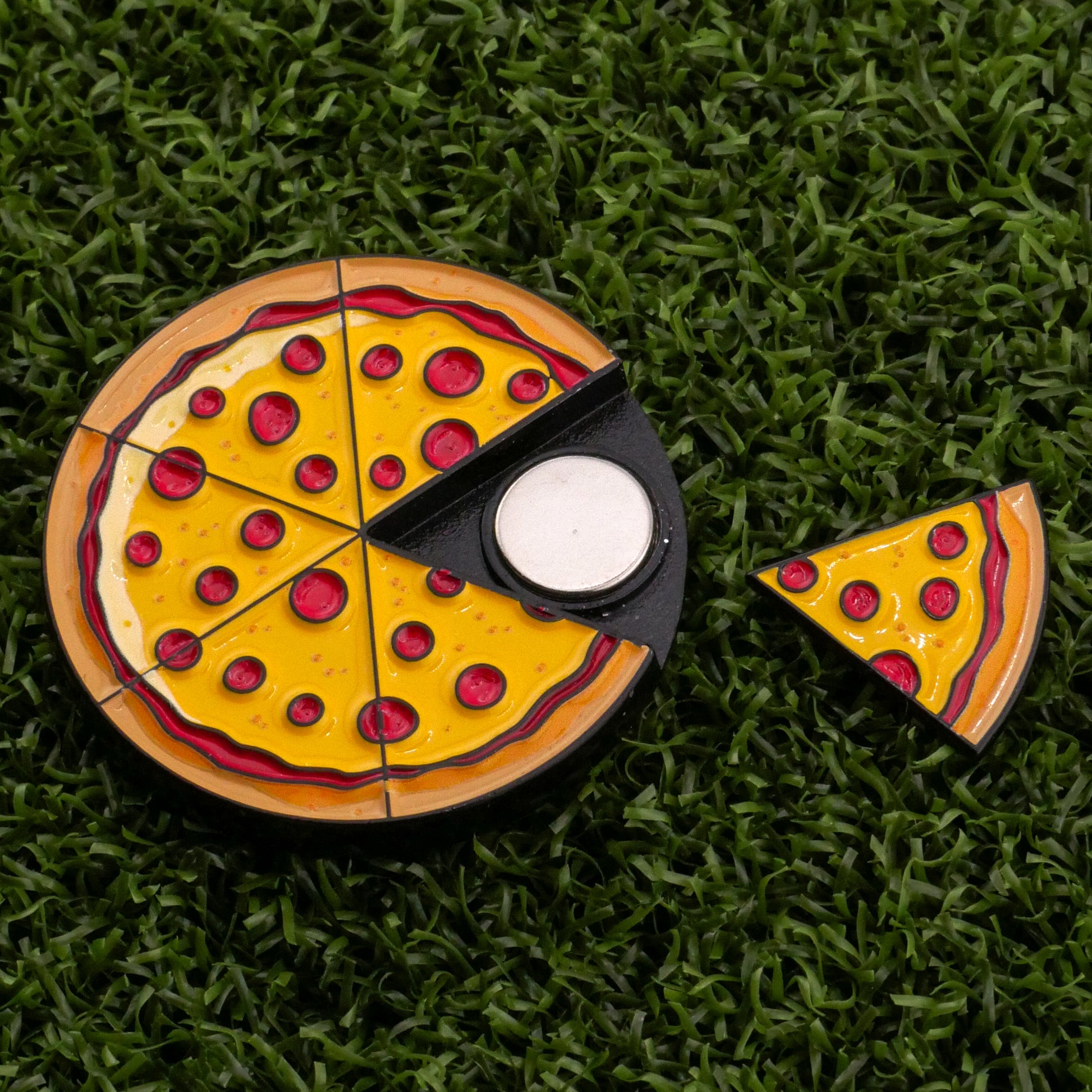 A Logie Joe's Golf Pepperoni Pizza "Slice" Premium Ball Marker, featuring a pizza with yellow cheese, red pepperoni, and one slice removed to reveal the silver ball marker, is displayed on a grassy surface.