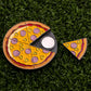Logie Joe's Golf Hawaiian Pizza "Slice" Premium Ball Marker is a pizza-shaped spinner with cheese and toppings, shown on grass. One removable slice serves as the ball marker, separated to reveal the central bearing.