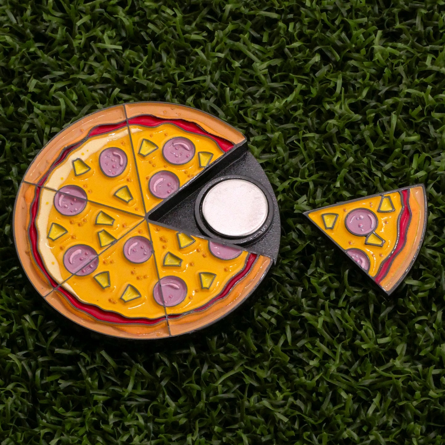 Logie Joe's Golf Hawaiian Pizza "Slice" Premium Ball Marker is a pizza-shaped spinner with cheese and toppings, shown on grass. One removable slice serves as the ball marker, separated to reveal the central bearing.