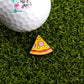 A Logie Joe's Golf ball sits on artificial grass next to the Hawaiian Pizza "Slice" Premium Ball Marker, a small, colorful removable marker shaped like a pizza slice.