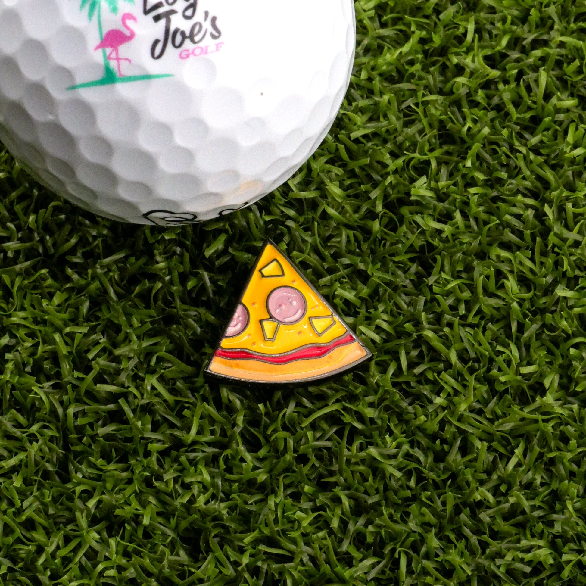A Logie Joe's Golf ball sits on artificial grass next to the Hawaiian Pizza "Slice" Premium Ball Marker, a small, colorful removable marker shaped like a pizza slice.