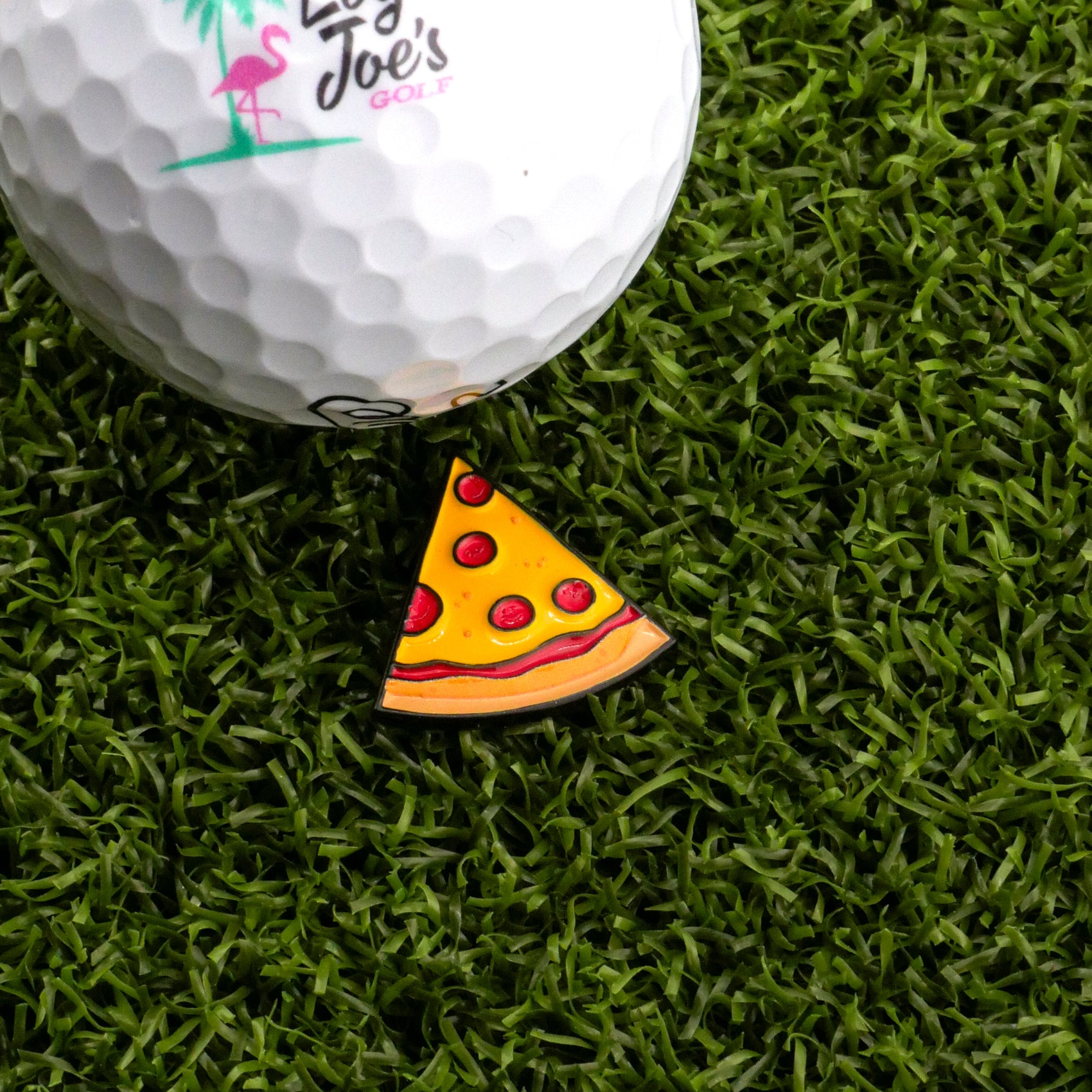 A golf ball with the Logie Joe's Golf logo sits on artificial grass next to the Pepperoni Pizza "Slice" Premium Ball Marker, shaped like a pizza slice.