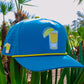 A vibrant blue Tequila and Lime Golf Hat by Logie Joe's Golf, featuring a lemon drink graphic, hangs on palm fronds—premium snapback cap with magnetic ball marker holder, marker included, and shot glass. Perfect for sunny golf days.