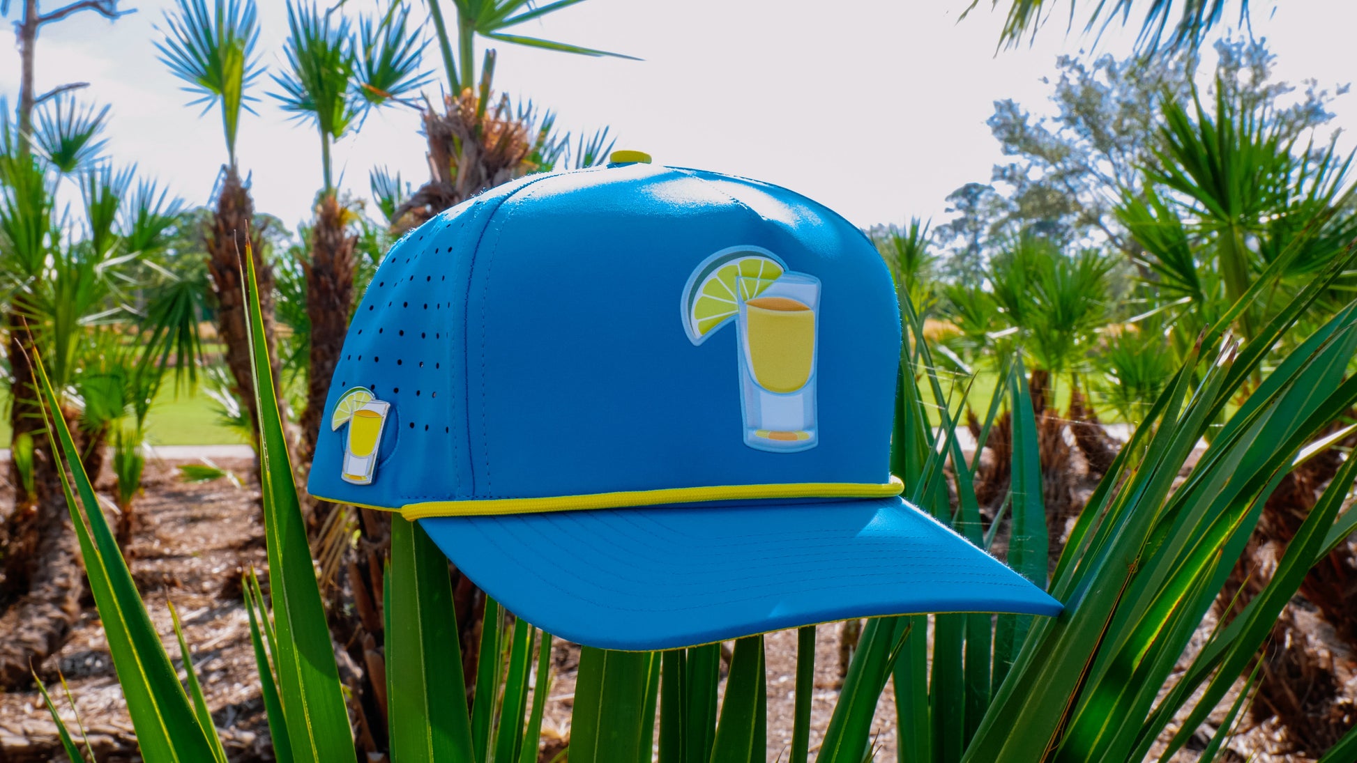 A vibrant blue Tequila and Lime Golf Hat by Logie Joe's Golf, featuring a lemon drink graphic, hangs on palm fronds—premium snapback cap with magnetic ball marker holder, marker included, and shot glass. Perfect for sunny golf days.