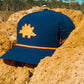 The Logie Joe's Golf Dino Nugget Snapback Hat + Matching Ball Marker, featuring an orange stegosaurus, sits on yellowish-brown rocks with trees and a clear blue sky in the background.