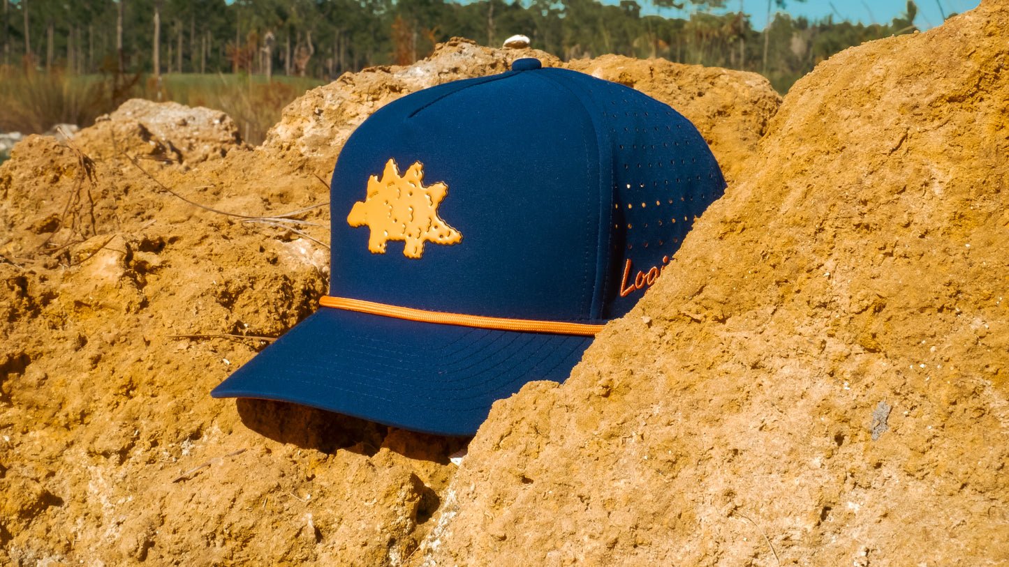 The Logie Joe's Golf Dino Nugget Snapback Hat + Matching Ball Marker, featuring an orange stegosaurus, sits on yellowish-brown rocks with trees and a clear blue sky in the background.