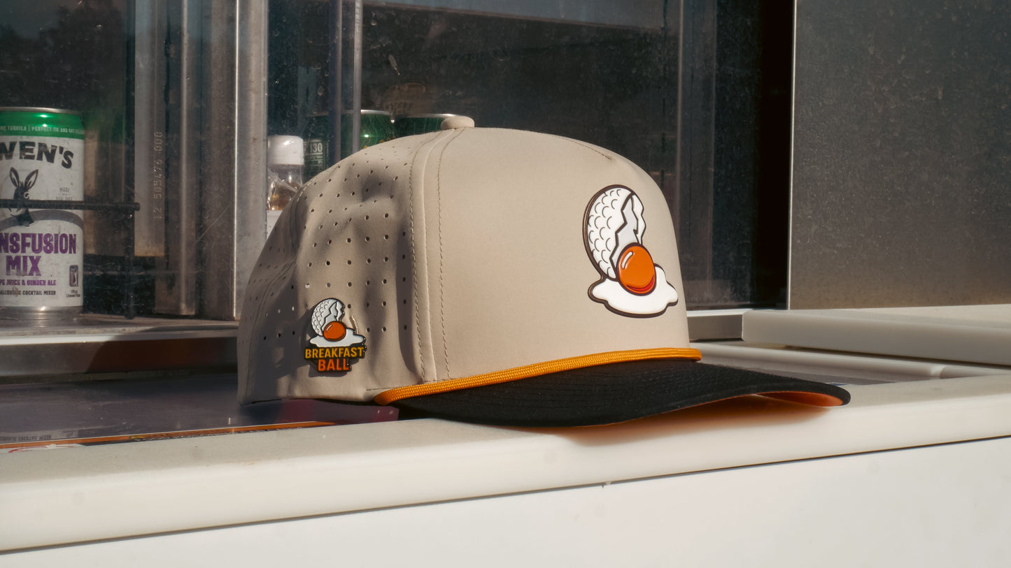 The Logie Joe's Golf Breakfast Ball Snapback Hat + Matching Ball Marker in beige and black features an embroidered egg-shaped golf ball with orange yolk, a “Breakfast Ball” side patch, and a built-in magnet for your marker.