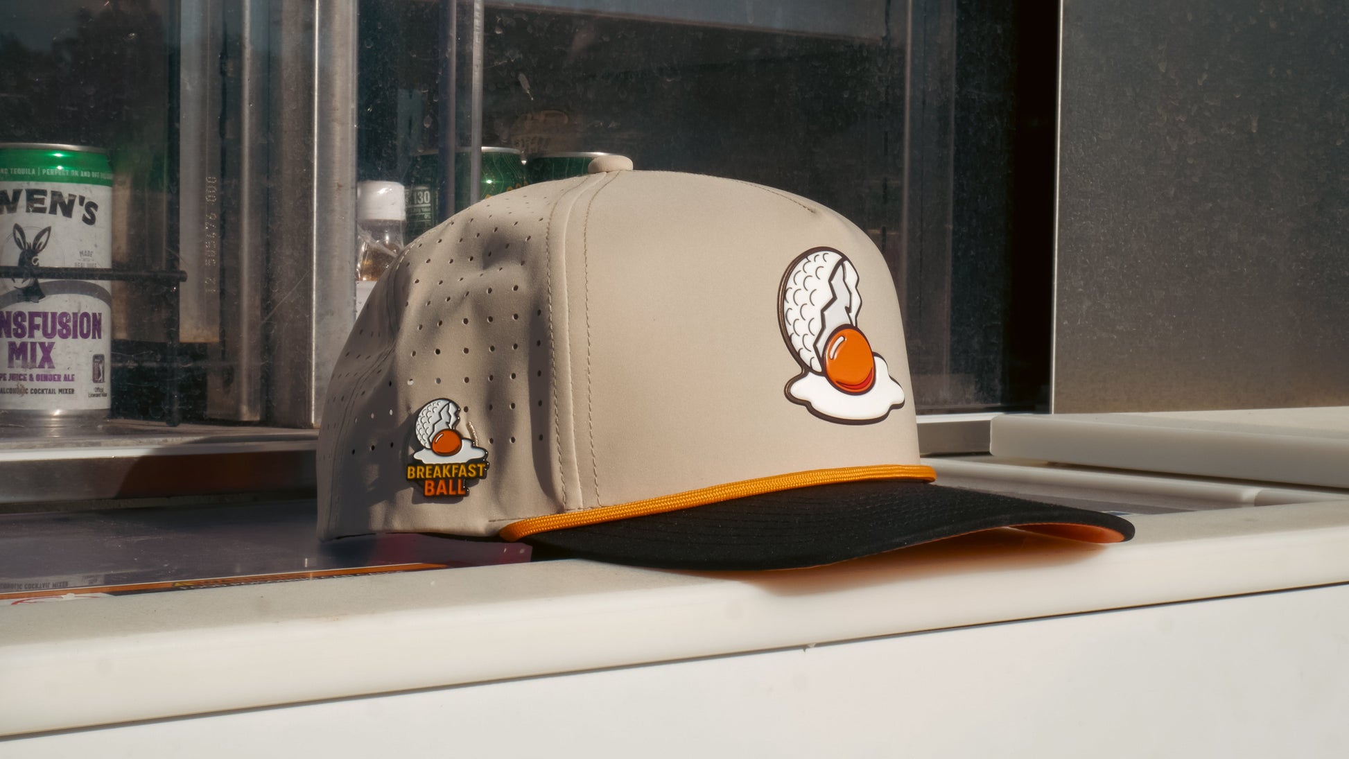 The Logie Joe's Golf Breakfast Ball Snapback Hat + Matching Ball Marker in beige and black features an embroidered egg-shaped golf ball with orange yolk, a “Breakfast Ball” side patch, and a built-in magnet for your marker.