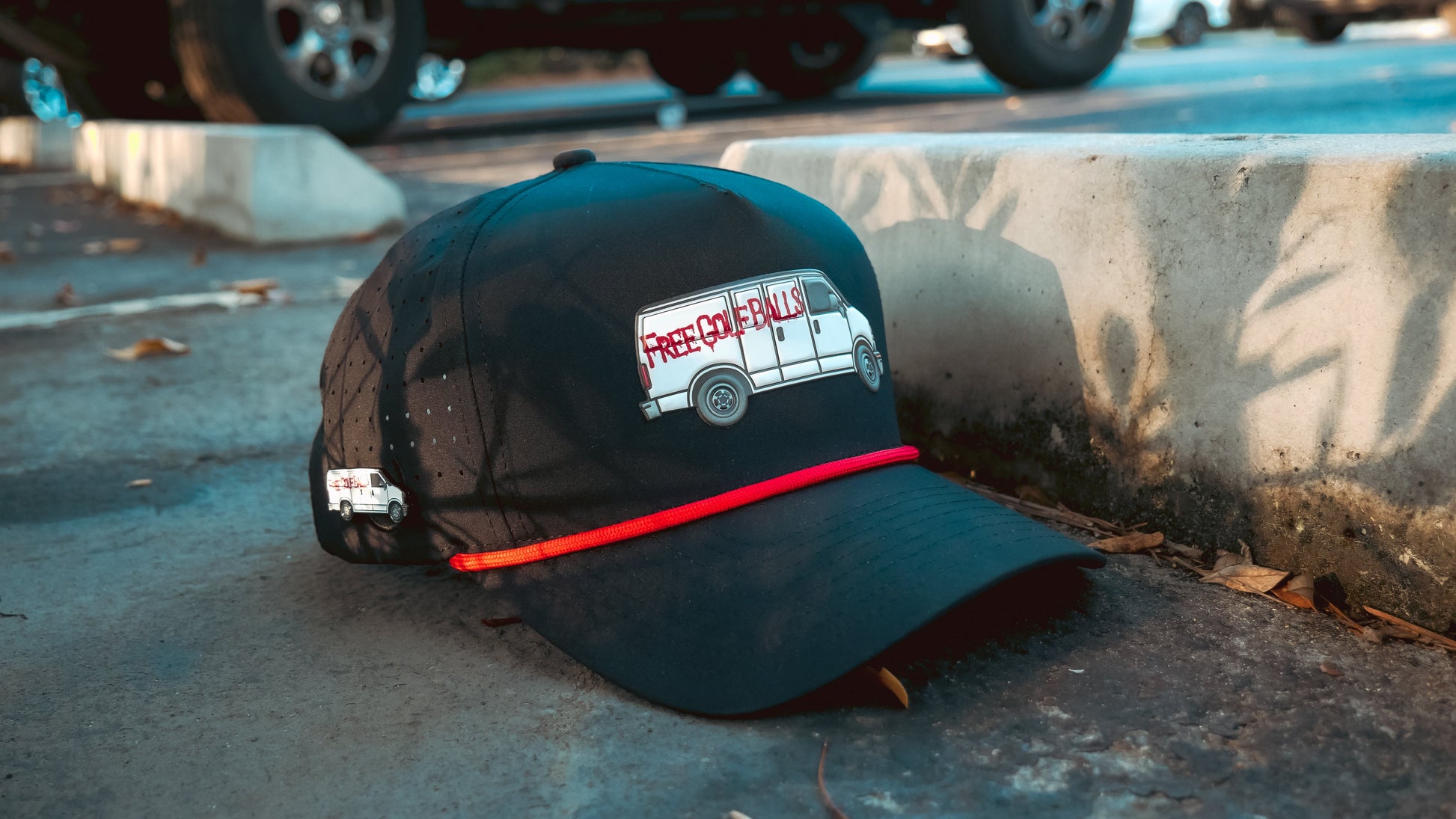 A Logie Joe’s Golf “Free Golf Balls Van” Snapback Hat with a matching ball marker sits on a concrete curb in a parking lot, with shadows and parked vehicles in the background.