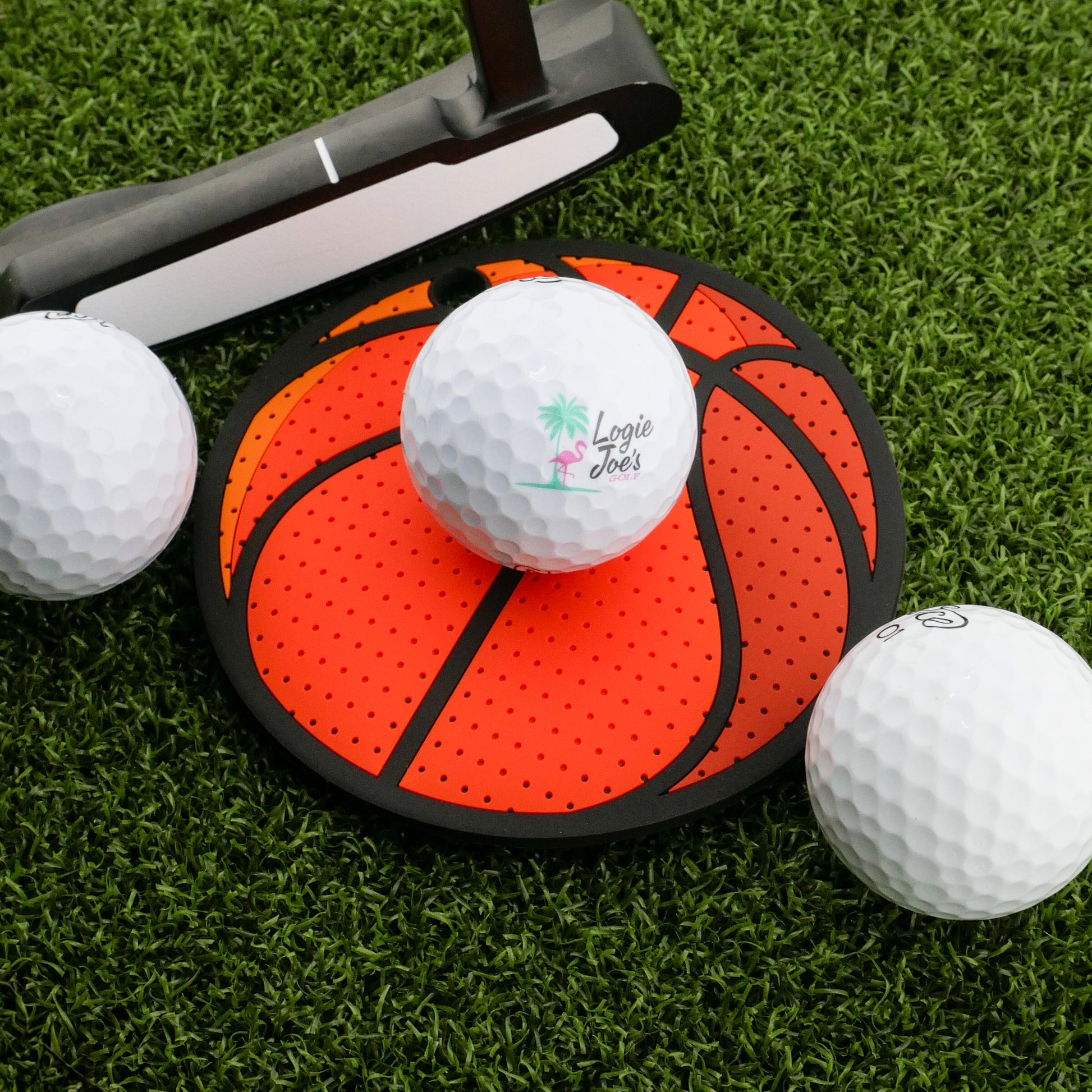 Three golf balls and a putter rest on artificial grass, with the Logie Joe's Golf Basketball Putting Disk—a disk styled like a basketball—at the center. Great for focused putting practice and stroke training.