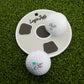 Two white golf balls, one labeled Logie Joe’s and the other Vice 01, sit on a round white Bottom of the Cup Putting Disk by Logie Joe’s Golf—ideal for practice—placed on green artificial turf.