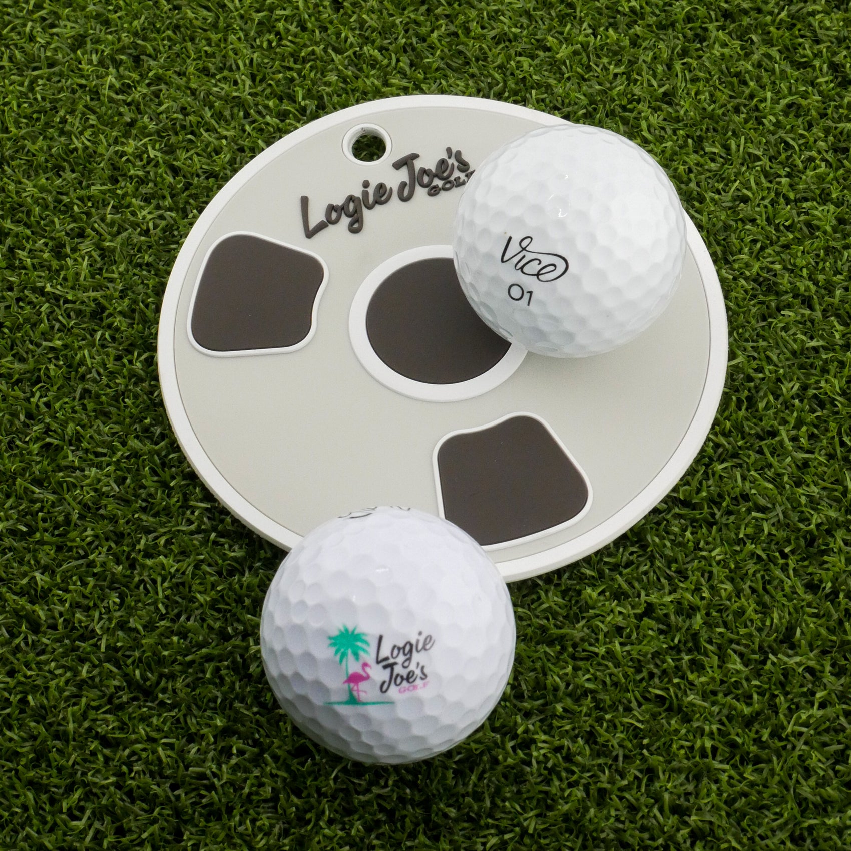 Two white golf balls, one labeled Logie Joe’s and the other Vice 01, sit on a round white Bottom of the Cup Putting Disk by Logie Joe’s Golf—ideal for practice—placed on green artificial turf.