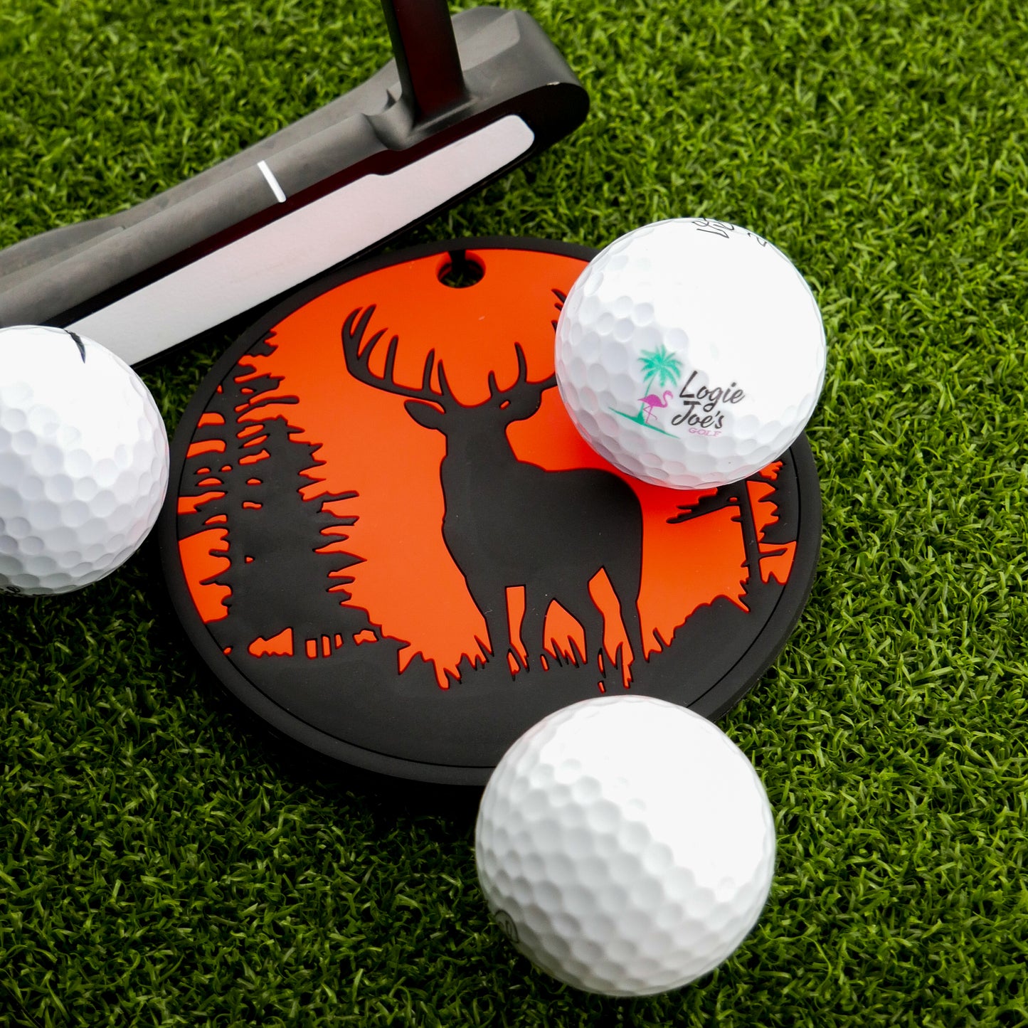 A Logie Joe's Golf Buck Hunter Putting Disk, a golf putter, and three golf balls are arranged on green artificial turf for focused putting practice.