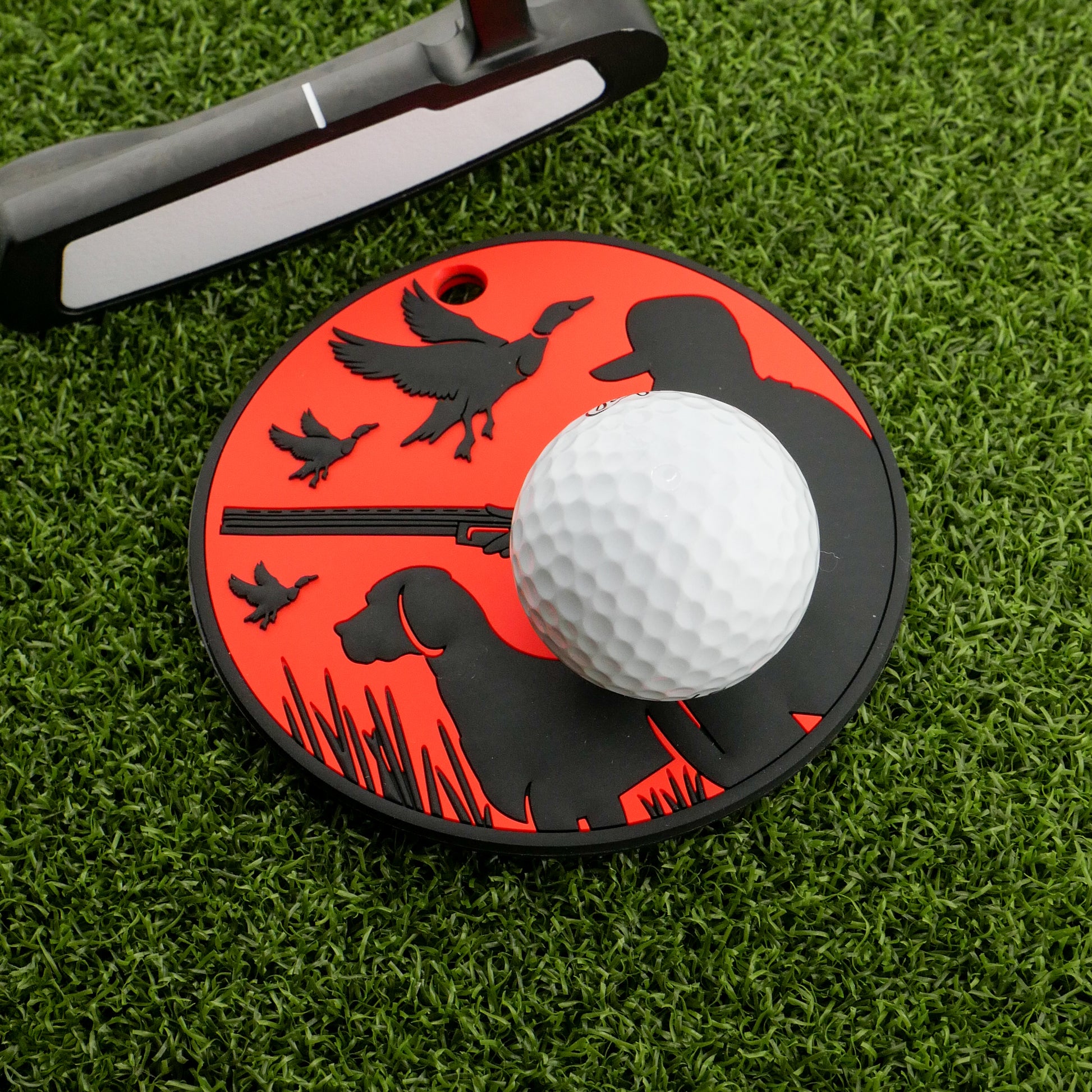 A white golf ball rests on Logie Joe's Golf Duck Hunter Putting Disk, which features black silhouettes of a hunter, dog, and birds. The red marker sits on artificial grass by a putter—ideal for stroke training or practice.