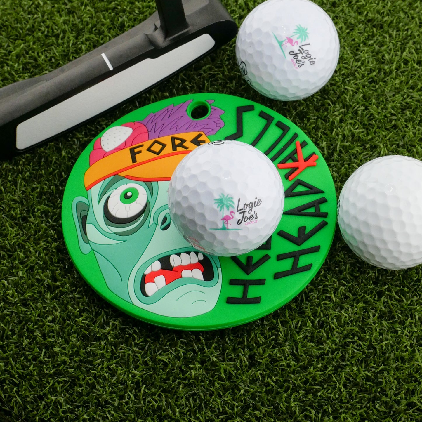 The Helmet Head Hills Putting Disk by Logie Joe's Golf features a cartoon zombie face and "FORE!"—a colorful marker perfect for stroke training, shown on green turf beside a putter and three Logie Joe's golf balls.