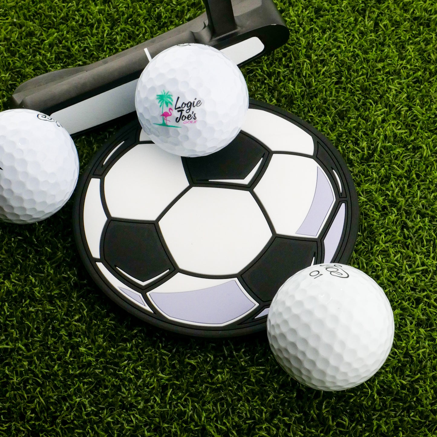 A Logie Joe's Golf Soccer Ball Putting Disk, a golf putter, and three white golf balls are set on artificial green grass for focused practice.