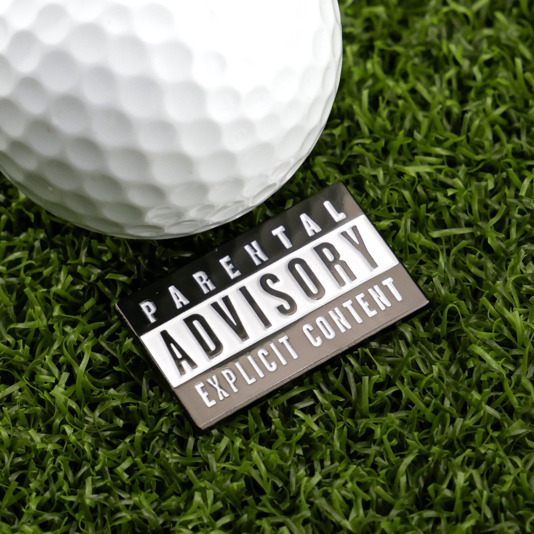 A white Parental Advisory golf ball by Logie Joe's Golf, featuring a black-and-white Explicit Content label, rests on artificial green grass—an eye-catching premium accessory for today’s golfer.