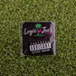 A black square sticker on green artificial grass shows Logie Joe's Golf, a pink flamingo, and "Parental Advisory"—ideal for fans of unique golf ball markers or premium accessories from Logie Joe's Golf.