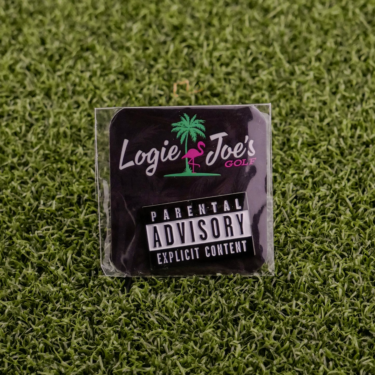 A black square sticker on green artificial grass shows Logie Joe's Golf, a pink flamingo, and "Parental Advisory"—ideal for fans of unique golf ball markers or premium accessories from Logie Joe's Golf.
