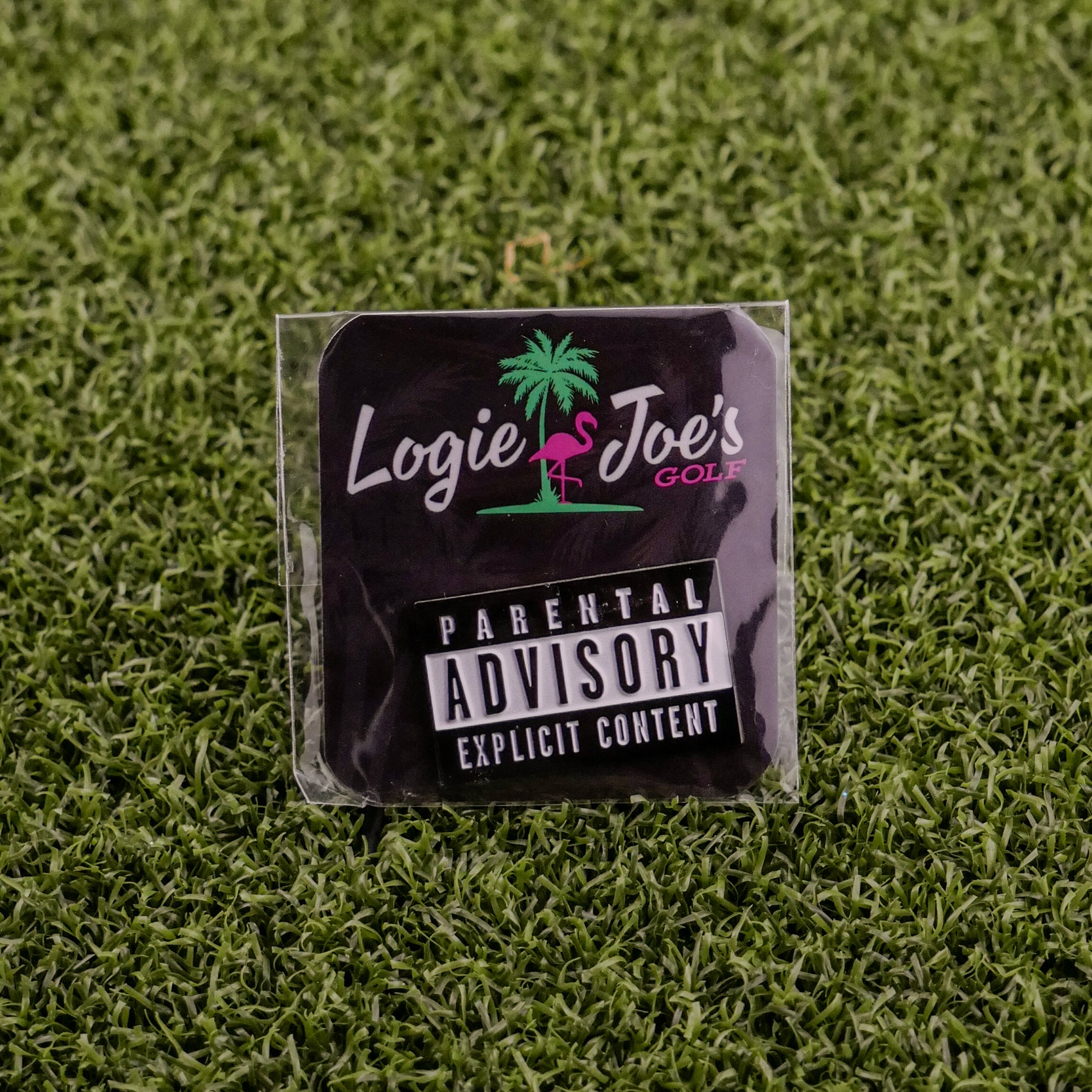 A black square sticker on green artificial grass shows Logie Joe's Golf, a pink flamingo, and "Parental Advisory"—ideal for fans of unique golf ball markers or premium accessories from Logie Joe's Golf.