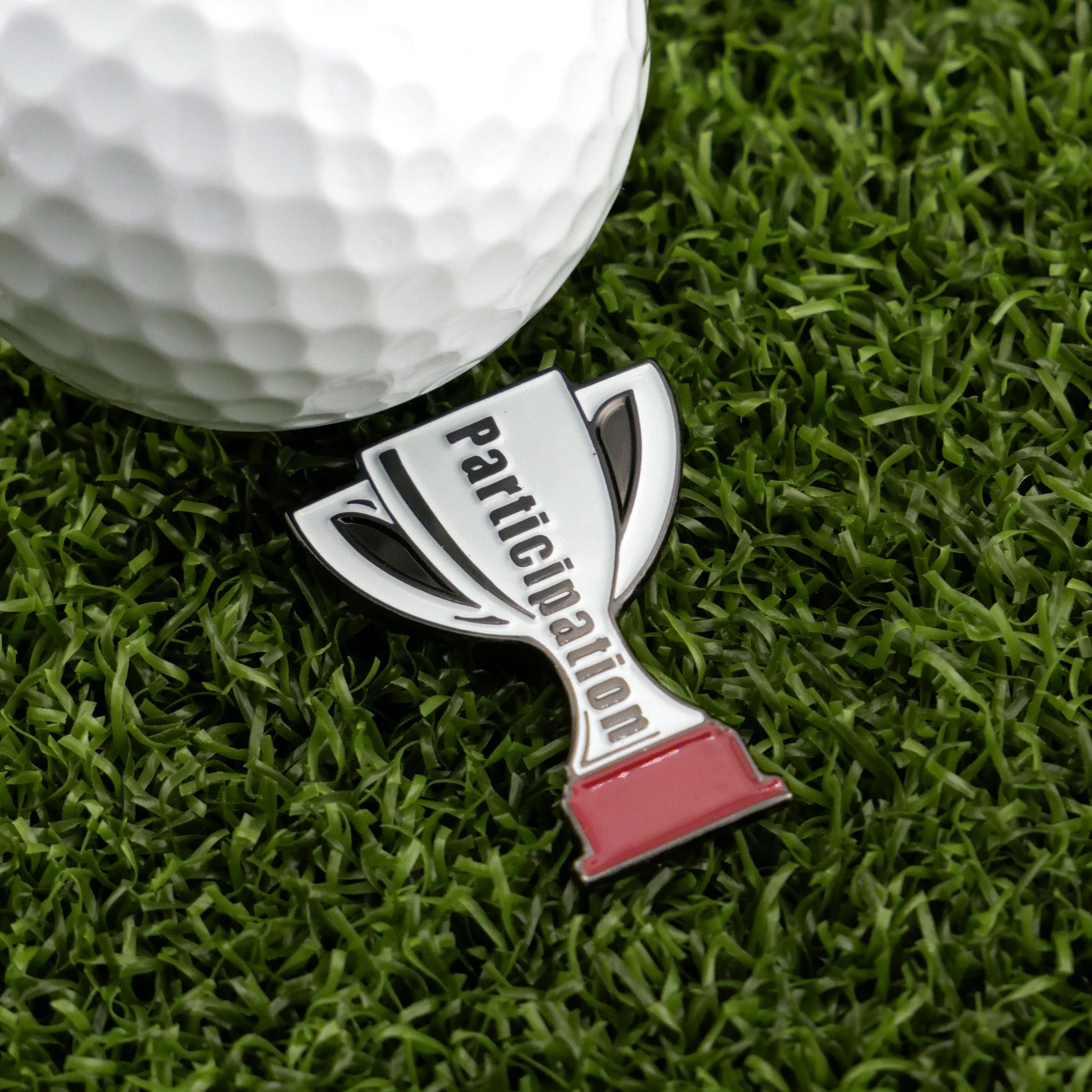 A close-up of Logie Joe's Golf Participation Trophy pin beside a golf ball on artificial grass, with premium golf accessories like ball markers adding to the scene.