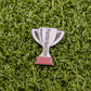 The Participation Trophy by Logie Joe's Golf is a trophy-shaped pin with "Participation" on it, set on green turf—an ideal playful golf ball marker or unique premium accessory for any golfer.