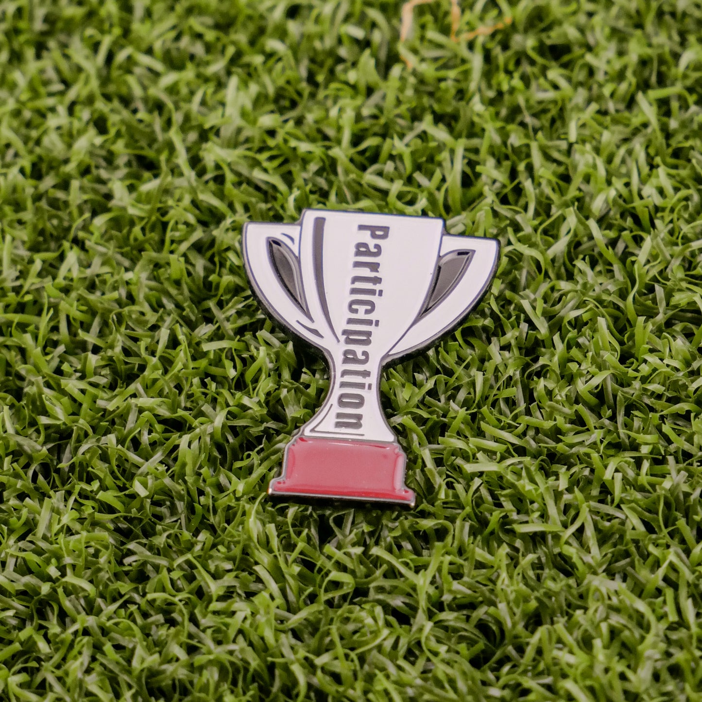 The Participation Trophy by Logie Joe's Golf is a trophy-shaped pin with "Participation" on it, set on green turf—an ideal playful golf ball marker or unique premium accessory for any golfer.