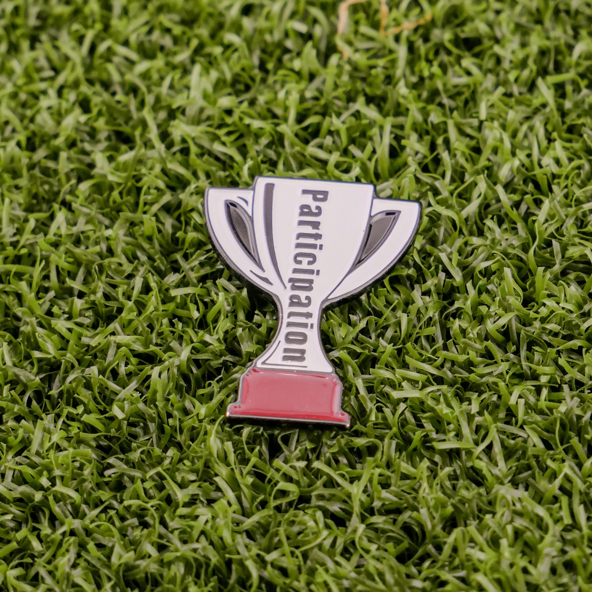 The Participation Trophy by Logie Joe's Golf is a trophy-shaped pin with "Participation" on it, set on green turf—an ideal playful golf ball marker or unique premium accessory for any golfer.