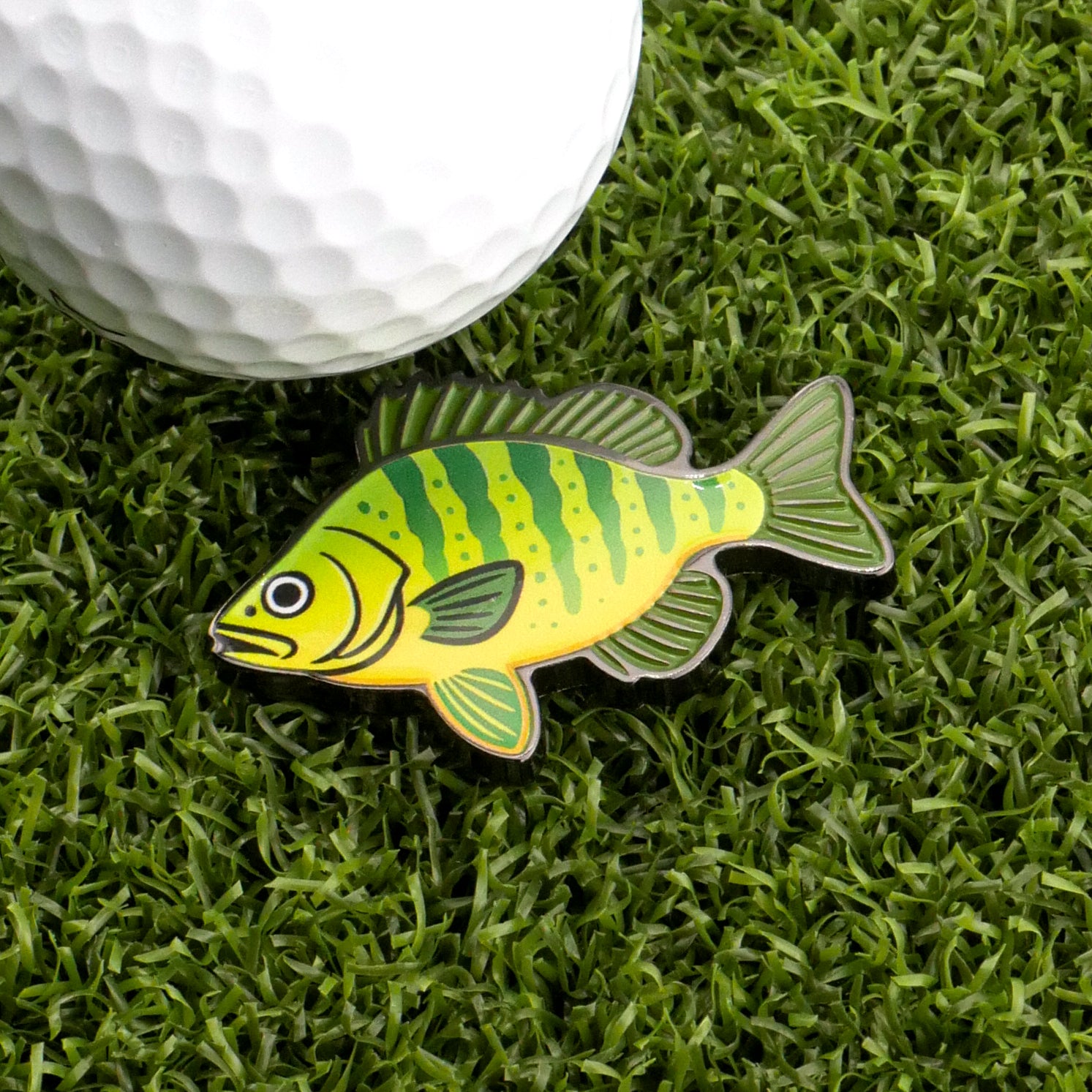 The Perch Ball Marker by Logie Joe's Golf, a cartoon fish-shaped accessory, sits on green artificial grass beside a white golf ball, adding playful style to your golf gear.